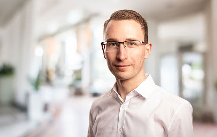 Eine junger lächelnder Mann mit weissem Hemd und schwarzer Brille.